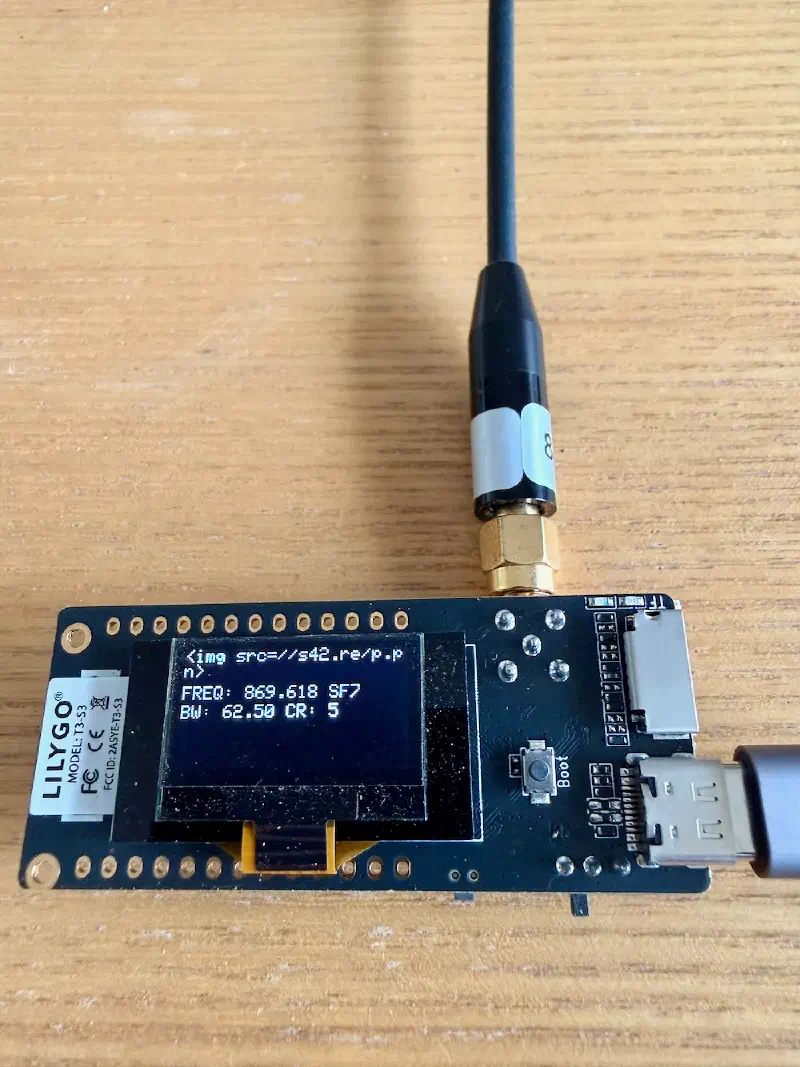 LILYGO T3-S3 LoRa node with antenna attached, OLED screen showing `<img src=//s42.re/p.pn>` as the node name.